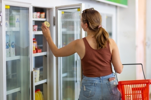 young woman with shopping basket taking yoghurt from shop refrigerator
